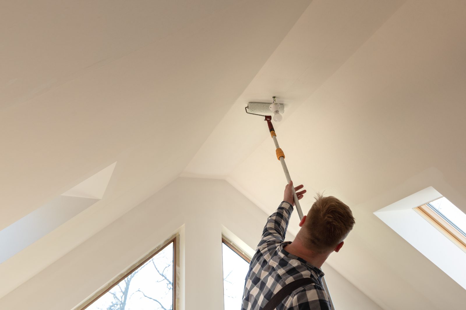 Worker painting ceiling with roller in attic room with skylights